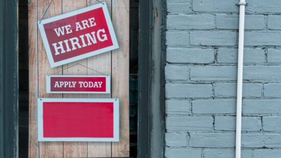"We Are Hiring" Sign in Window of Brick Building - A red and white sign in a window of a brick building reads We Are Hiring with a smaller sign that says Apply Today.